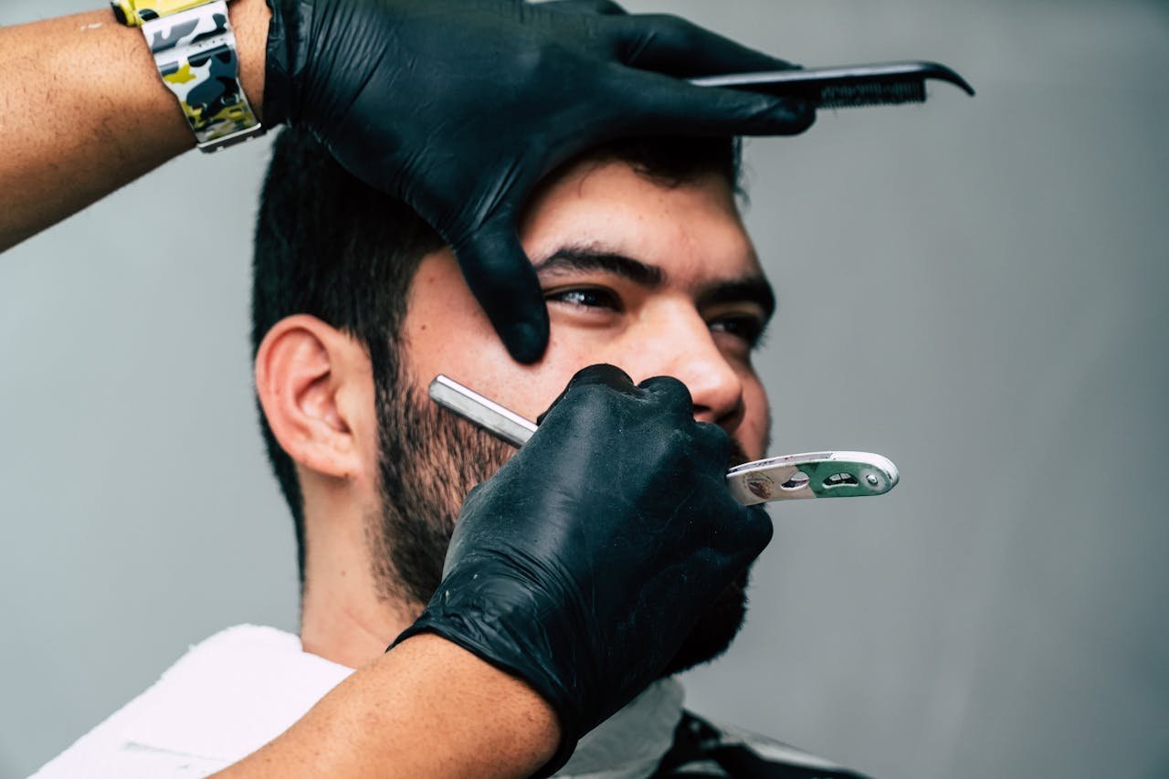 Servicios. Close-up of a barber shaving a mans beard with precision in a contemporary barbershop setting.